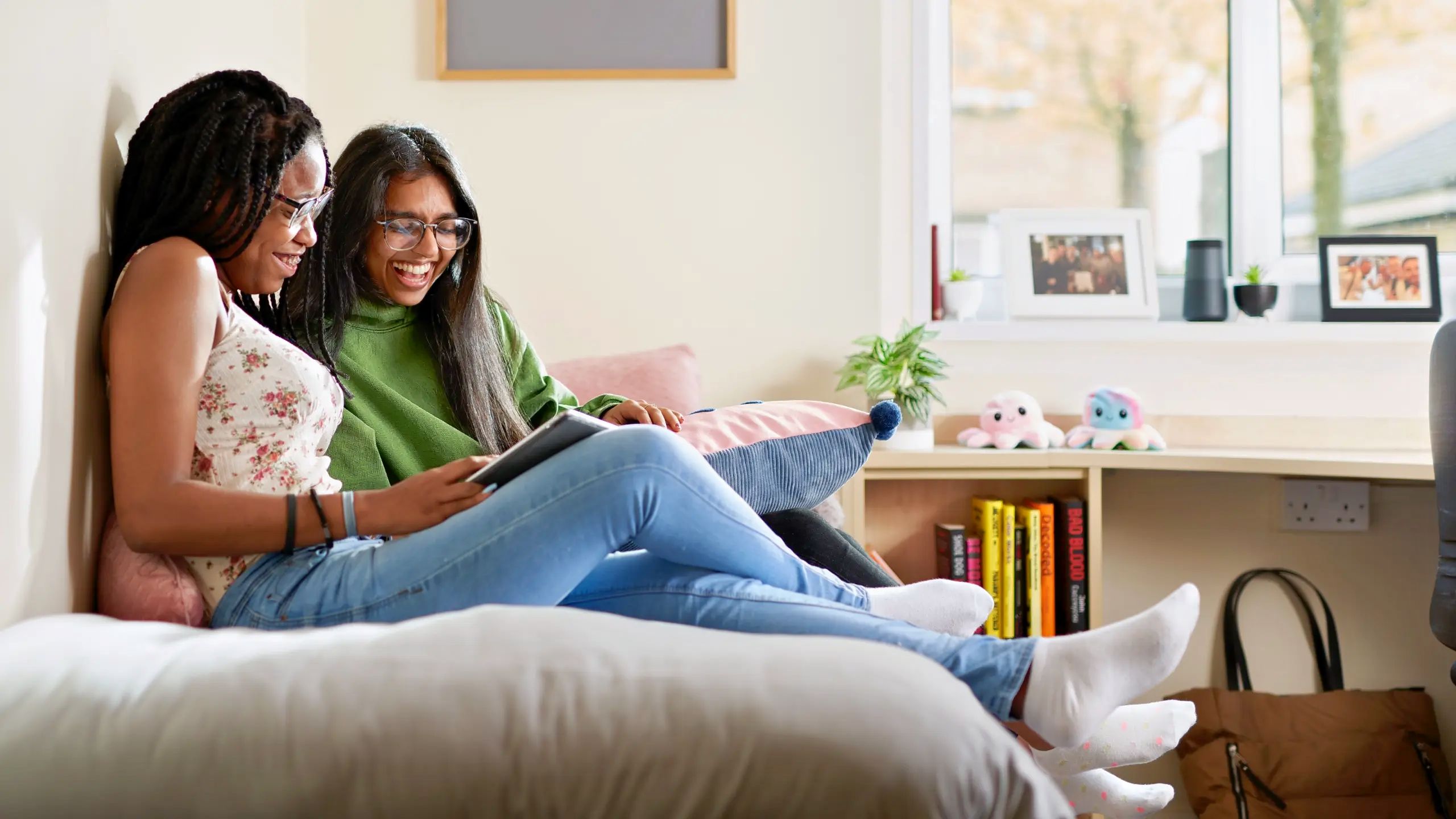 Two students sit on a bed reading and laughing.