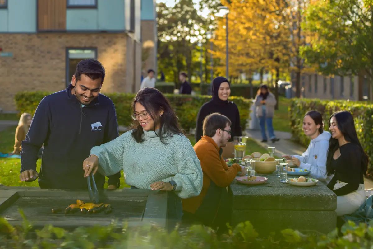Two students are grilling food on a barbecue while more sit behind them at a picnic table, eating.