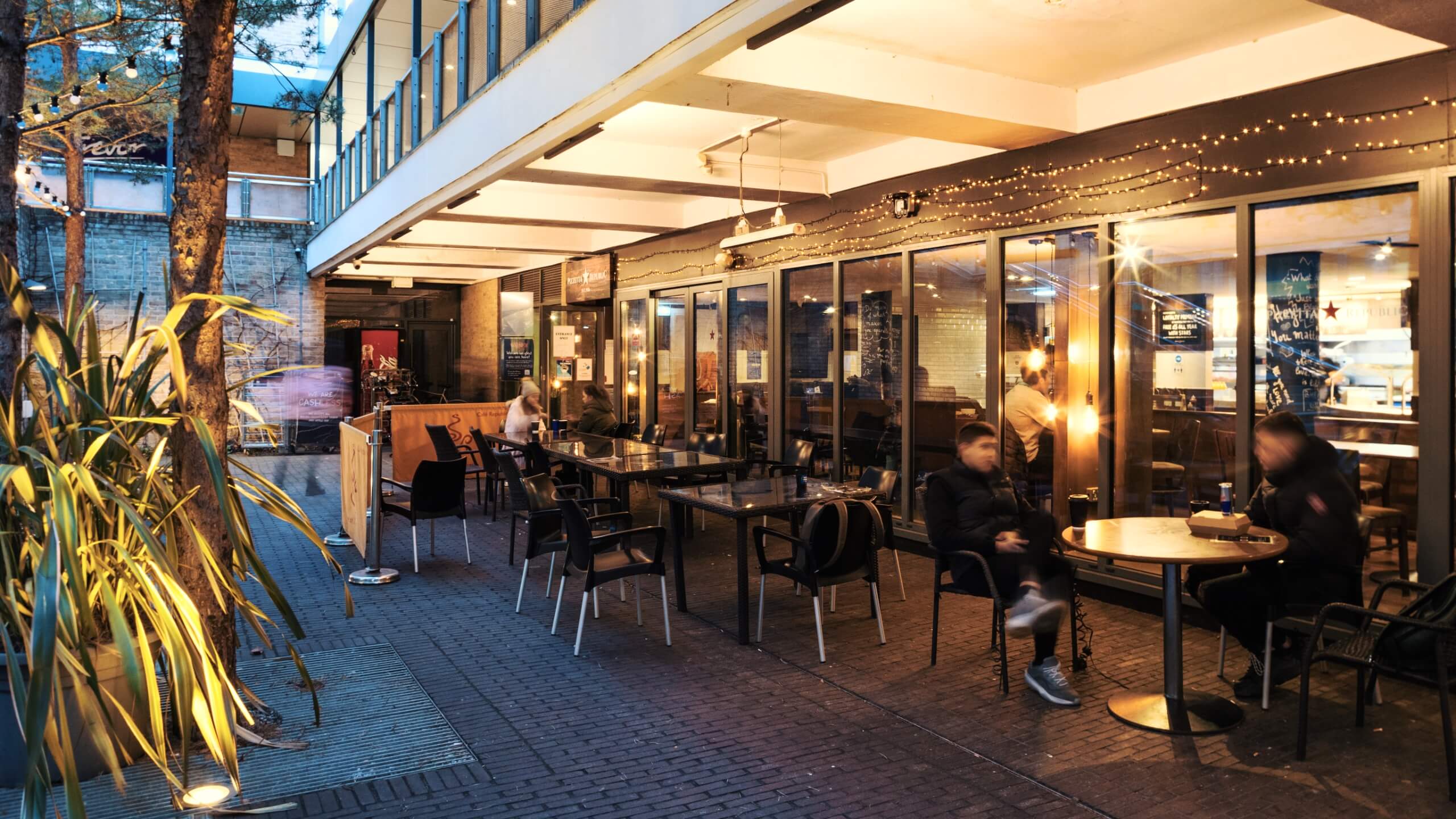 People sit outside a restaurant at dusk, with decorative lighting around them.