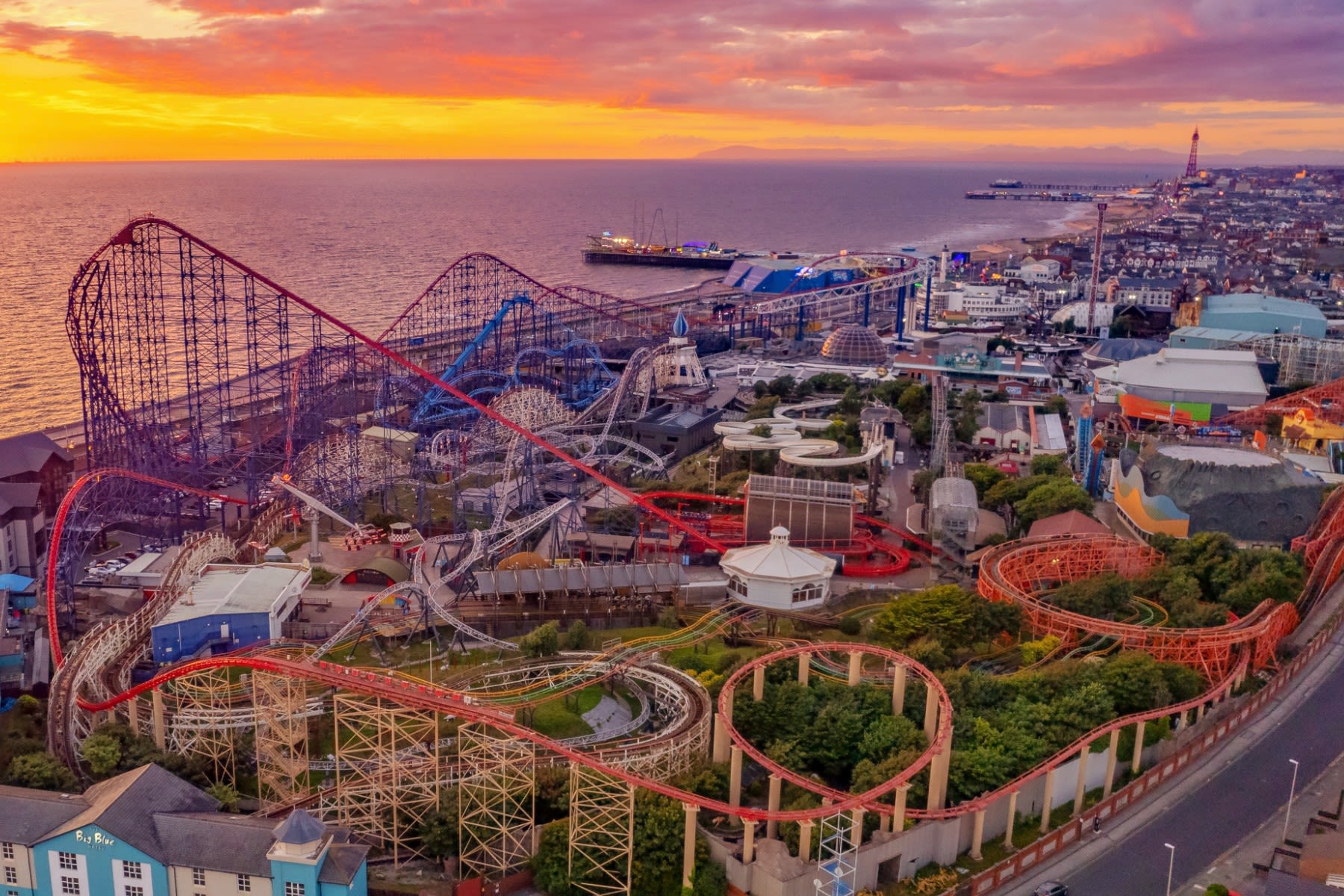 Blackpool Pleasure Beach at sunset, with Blackpool Tower and the Lake District hills in the distance.