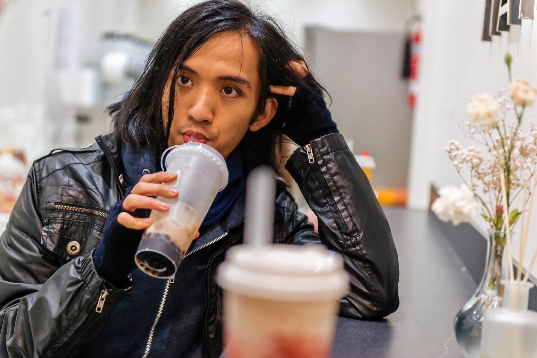 A male student in a leather jacket slurps on a boba tea in a cafe.