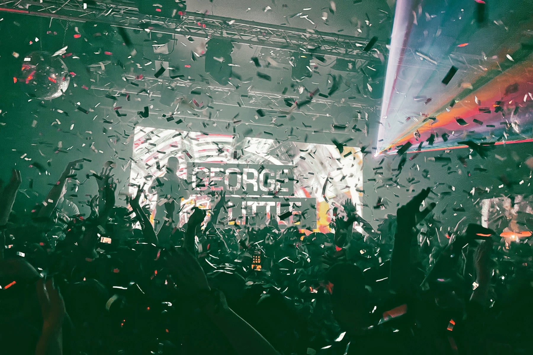 Interior of a crowded nightclub, with flashing lights. Confetti falls from the ceiling onto the crowds below.