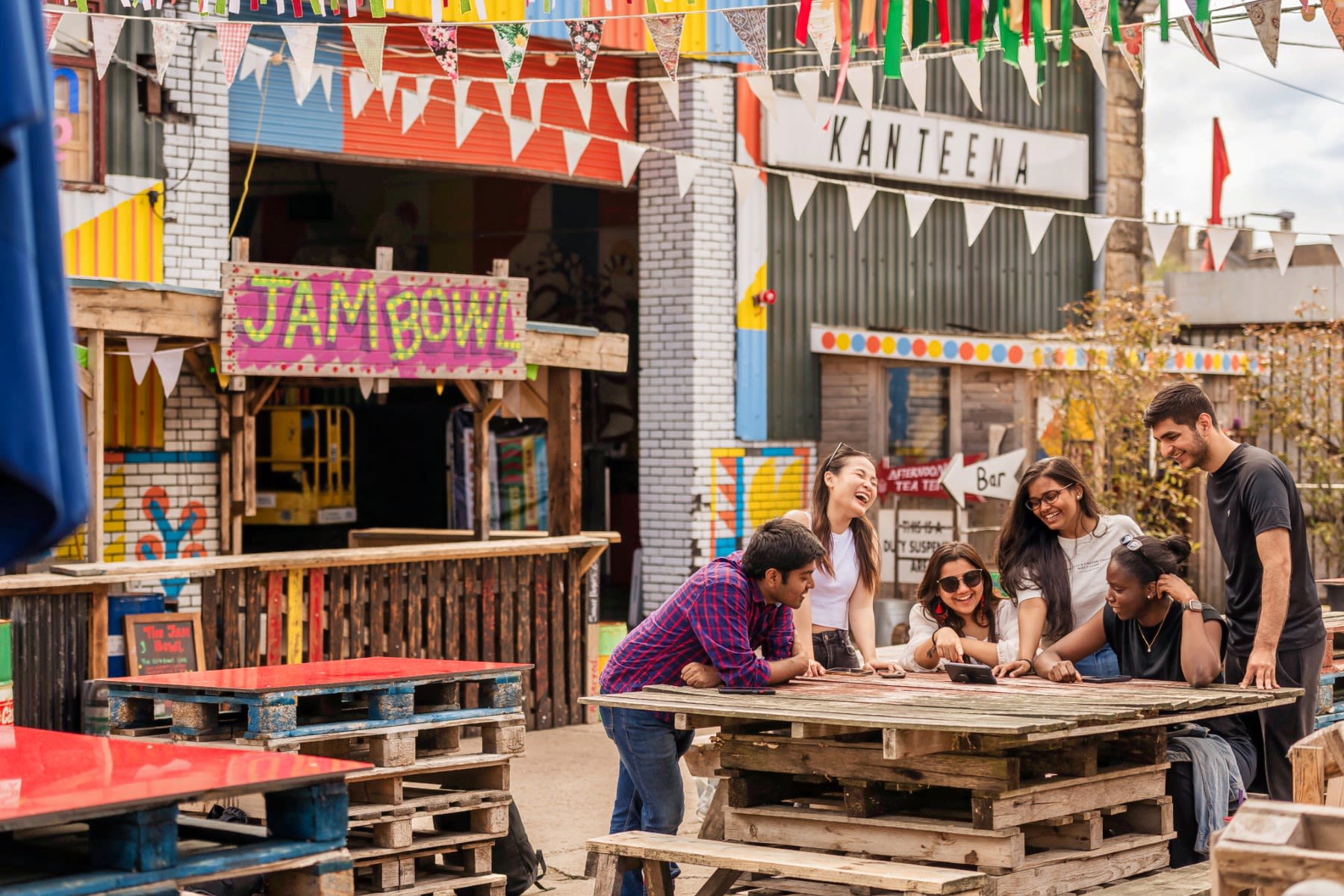 Students sit outside Kanteena, laughing and pointing at a mobile phone.