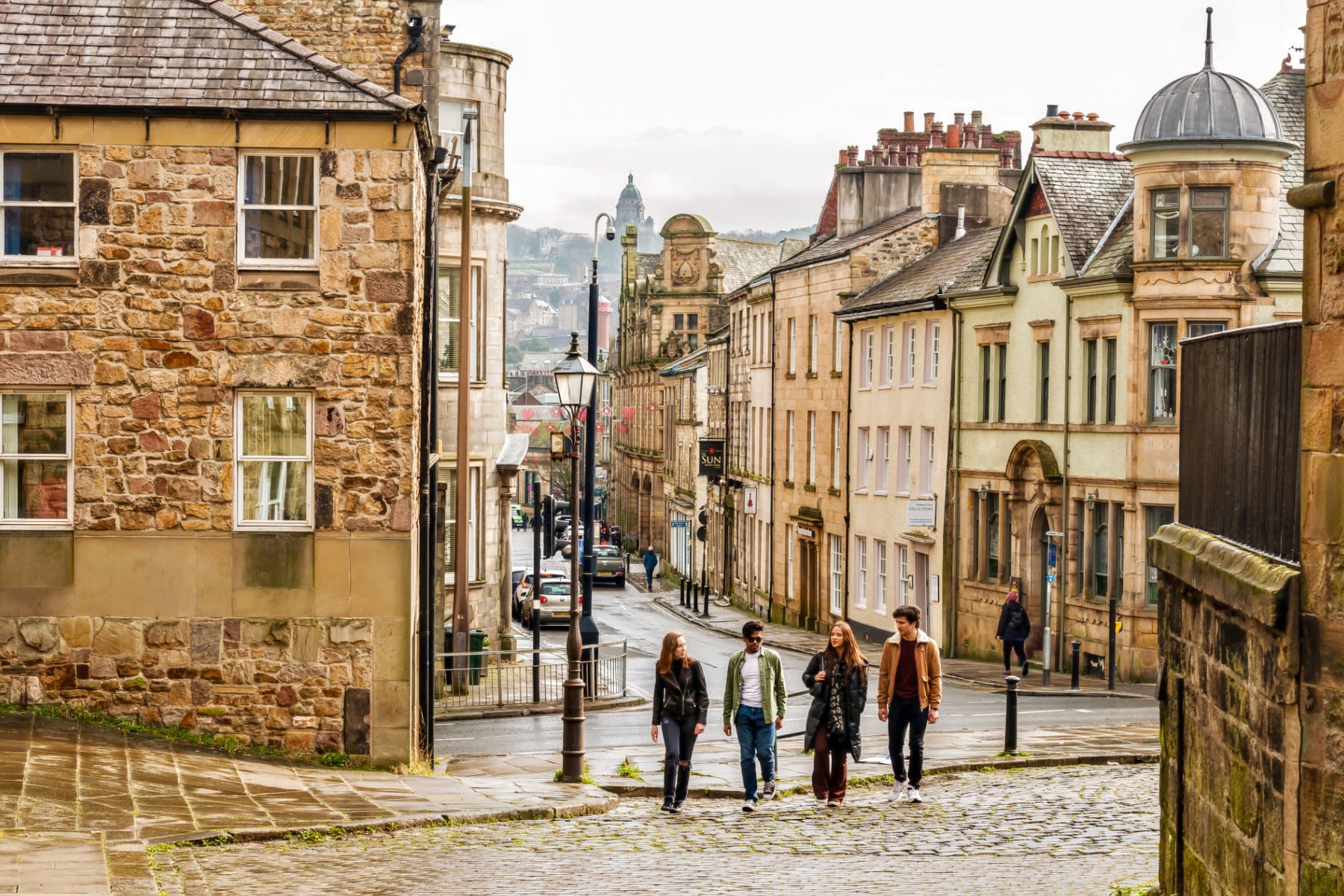 Four students walk on cobbled streets surrounded by historic buildings. The Ashton Memorial is on a hill top in the background.