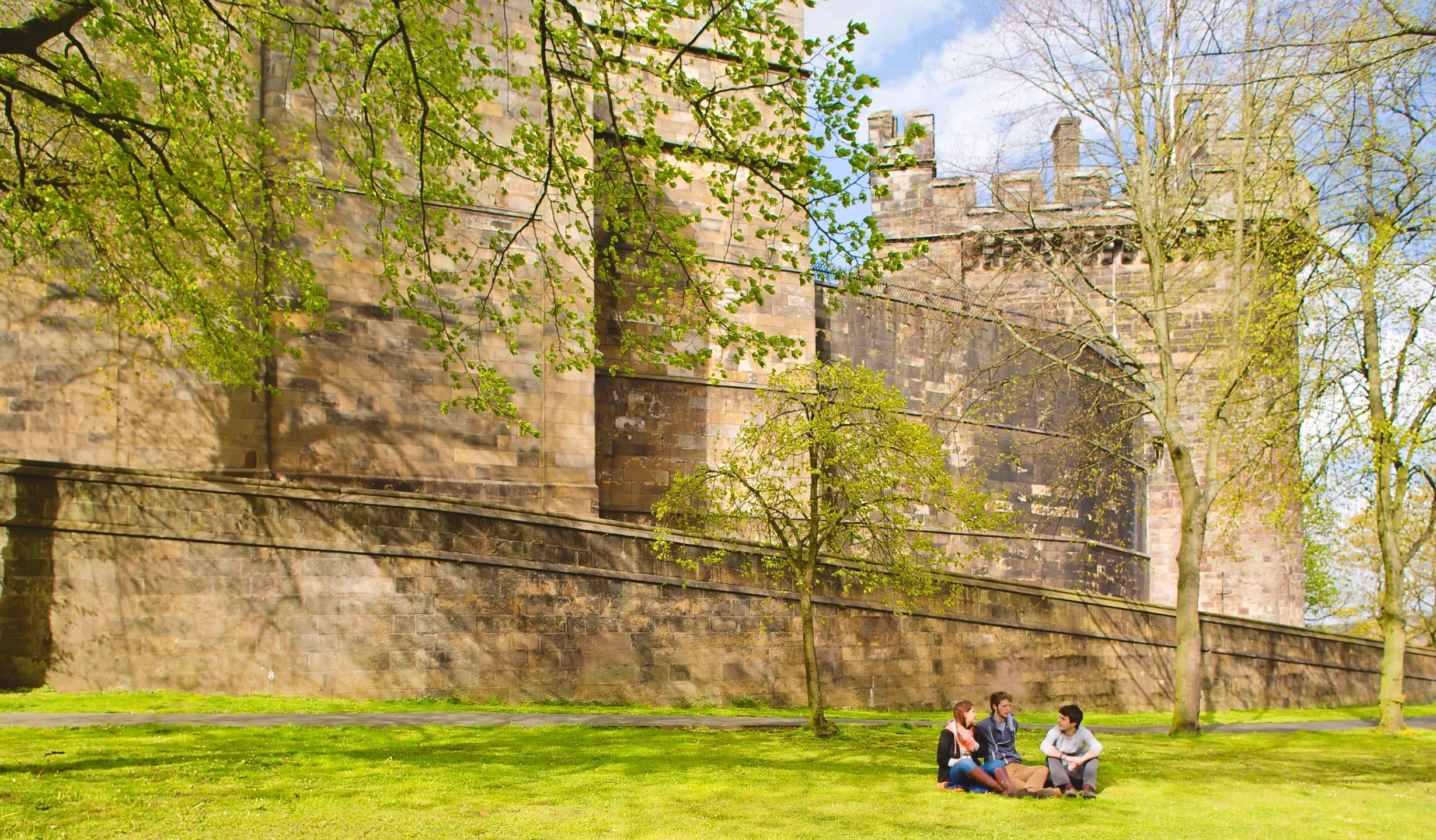 Three students sit in the sun on a patch of grass outside the walls of Lancaster Castle.
